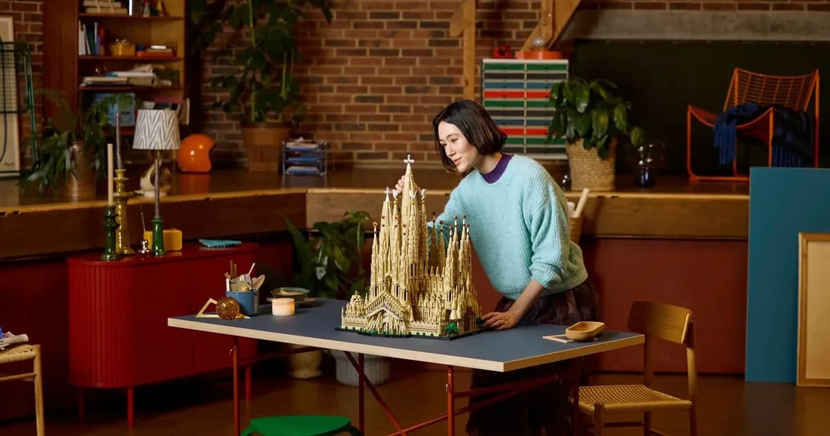A woman leaning over a tall tan LEGO Sagrada Família model on a blue table in a brick-walled living room