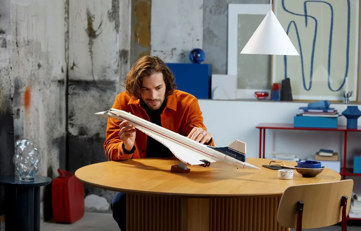 A man fitting the white LEGO Concorde airliner model on its stand on a round wooden table