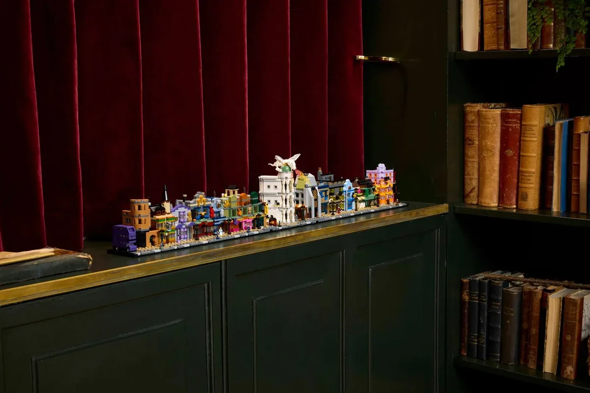 A long row of small colourful LEGO Diagon Alley shop buildings on a dark green cabinet beside a bookshelf