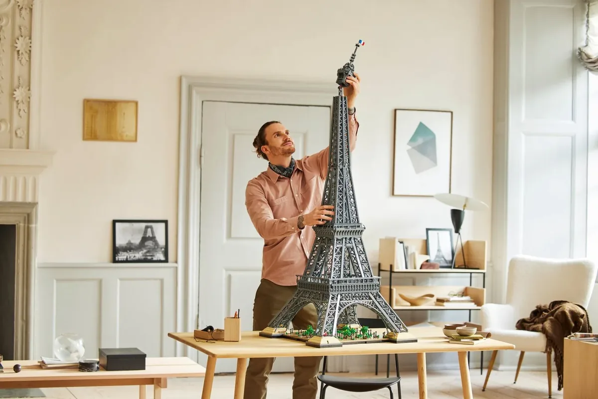 A man placing the top section on a tall grey LEGO Eiffel Tower model on a wooden table in a bright room
