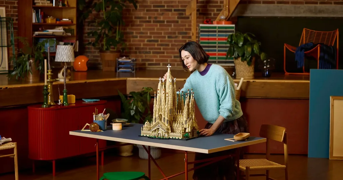 A woman leaning over a tall tan LEGO Sagrada Família model on a blue table in a brick-walled living room