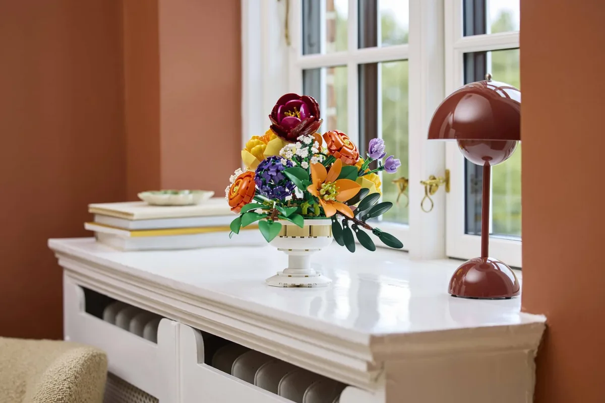 The LEGO Flower Arrangement 10345 on a white sideboard by a window, next to a dark red lamp and a stack of books