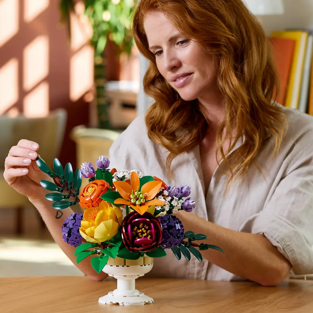 A woman adjusting the stems of the LEGO Flower Arrangement 10345, with orange lilies, red peonies and purple hydrangeas in a white chalice
