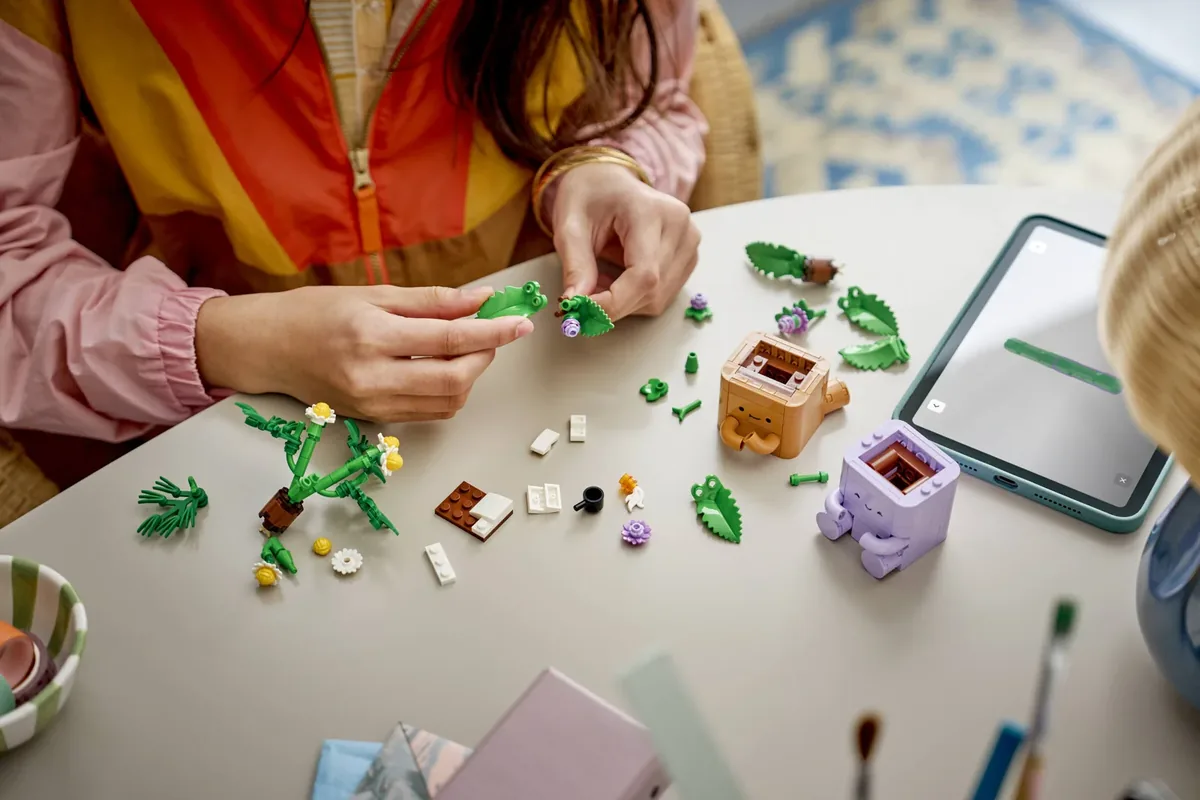 Two hands assembling LEGO Cozy Plants on a table, with green leaf pieces, flower parts and the two half-built pots laid out next to a tablet