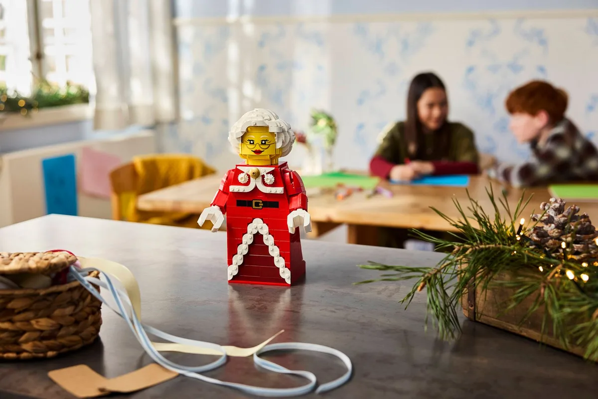 The LEGO Up-Scaled Mrs. Claus figure in a red coat standing on a table next to pine branches and a basket