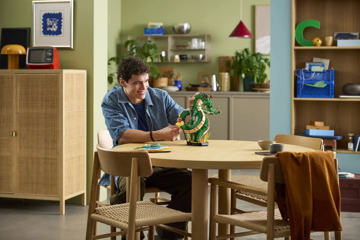 A man in a denim shirt sits at a round wooden table with the finished Shenron model, shelves and a cabinet behind him