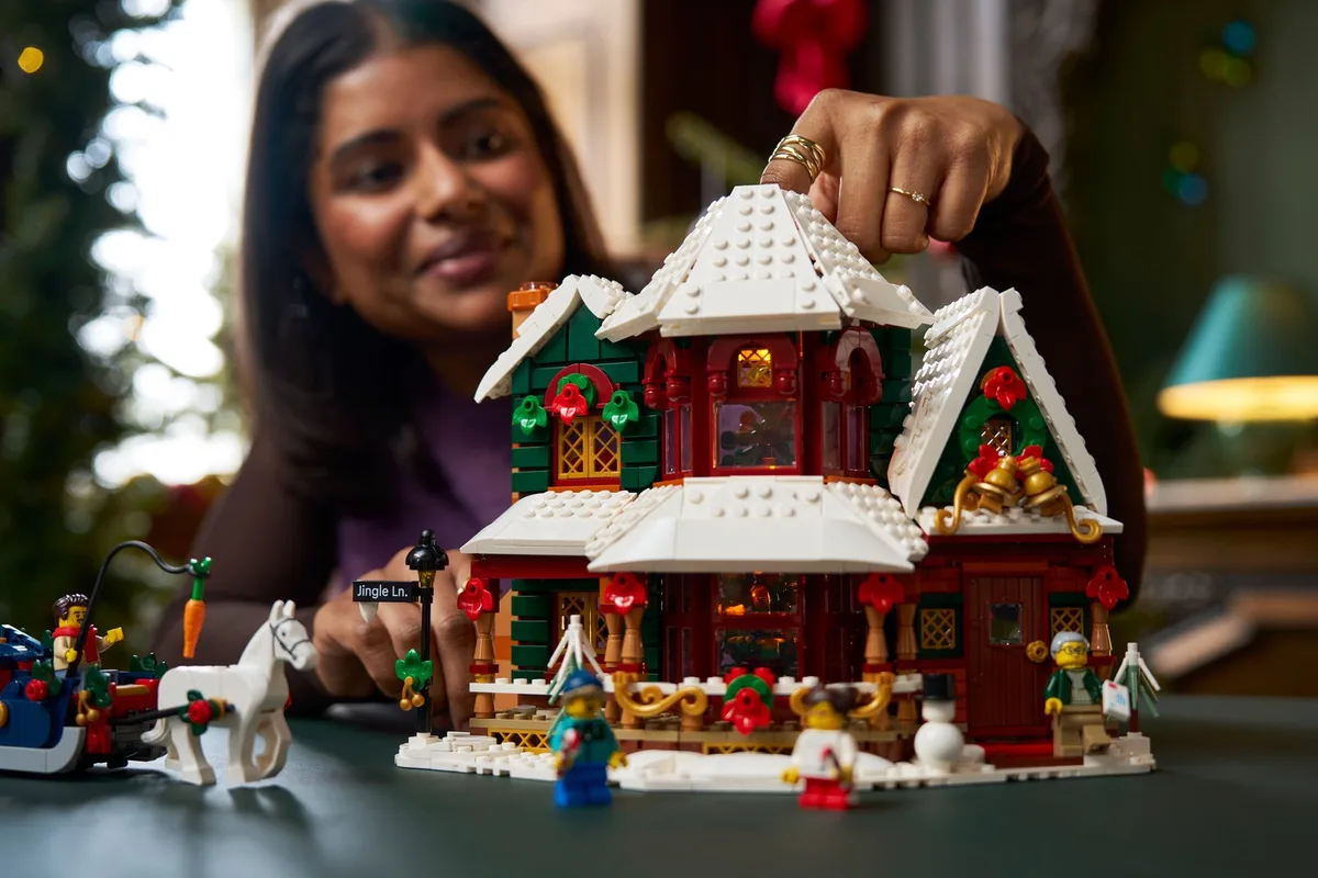 A woman placing the snowy roof on the LEGO Holiday House 11387, with the sleigh and white horse in front