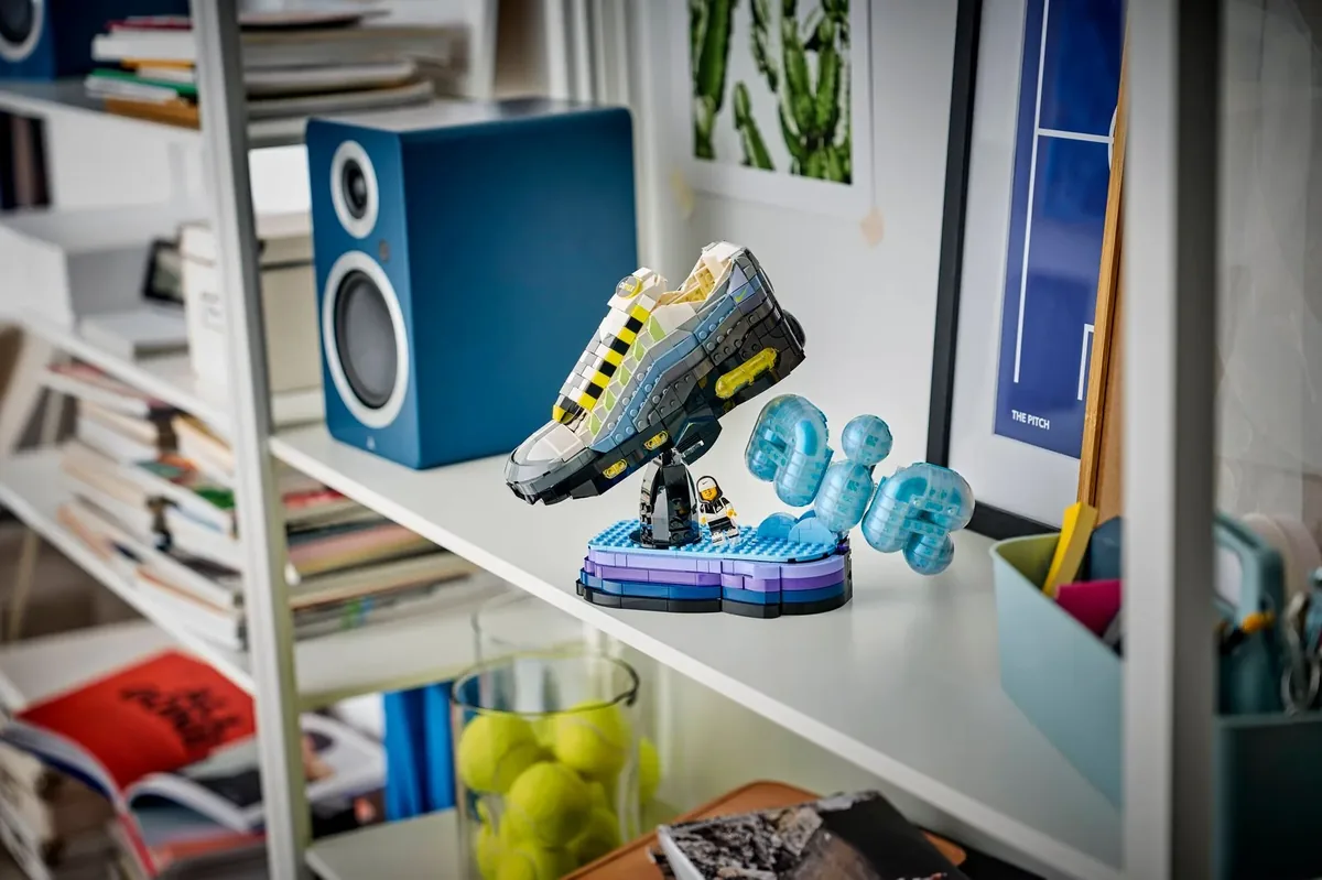 LEGO Nike Air Max 95 sneaker in grey, yellow and black on a blue stand with an Air logo, on a white shelf beside a speaker and books
