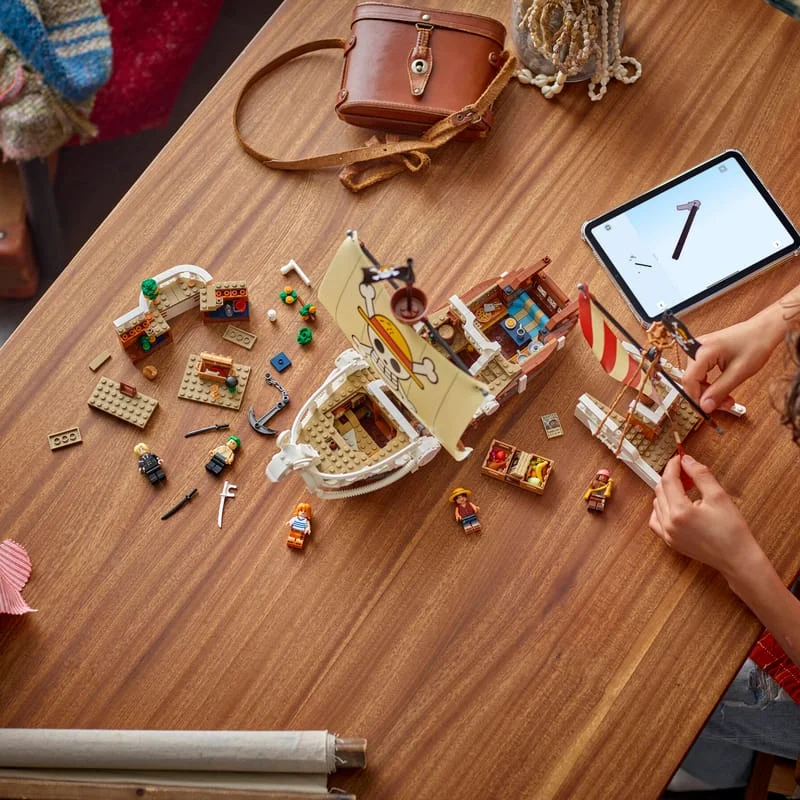 LEGO ONE PIECE Going Merry pirate ship built on a wooden table, with minifigures and loose parts laid out beside a tablet and a leather bag