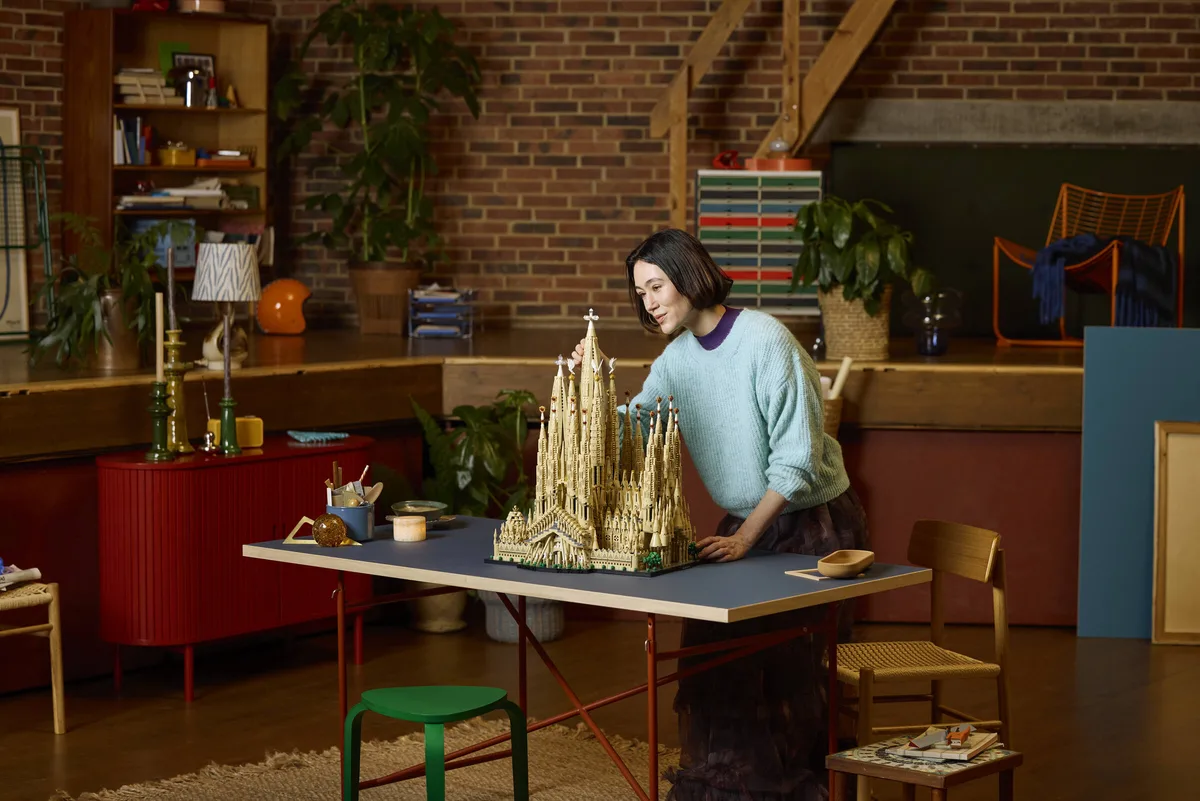 A woman in a pale blue sweater leans over the finished LEGO Sagrada Família model on a long table in a brick-walled room