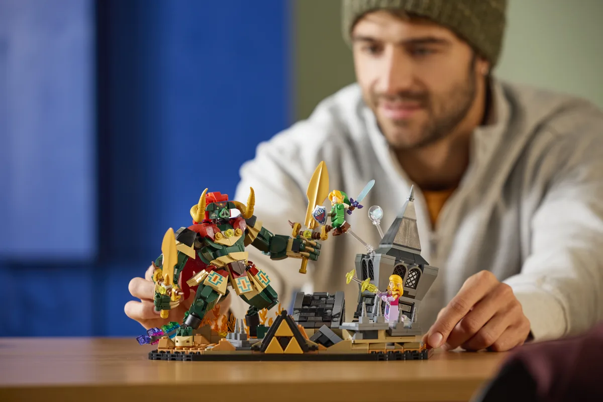 A man in a beanie arranges the LEGO Ocarina of Time Final Battle scene, a horned Ganon figure facing Link and a stone tower, on a wooden table