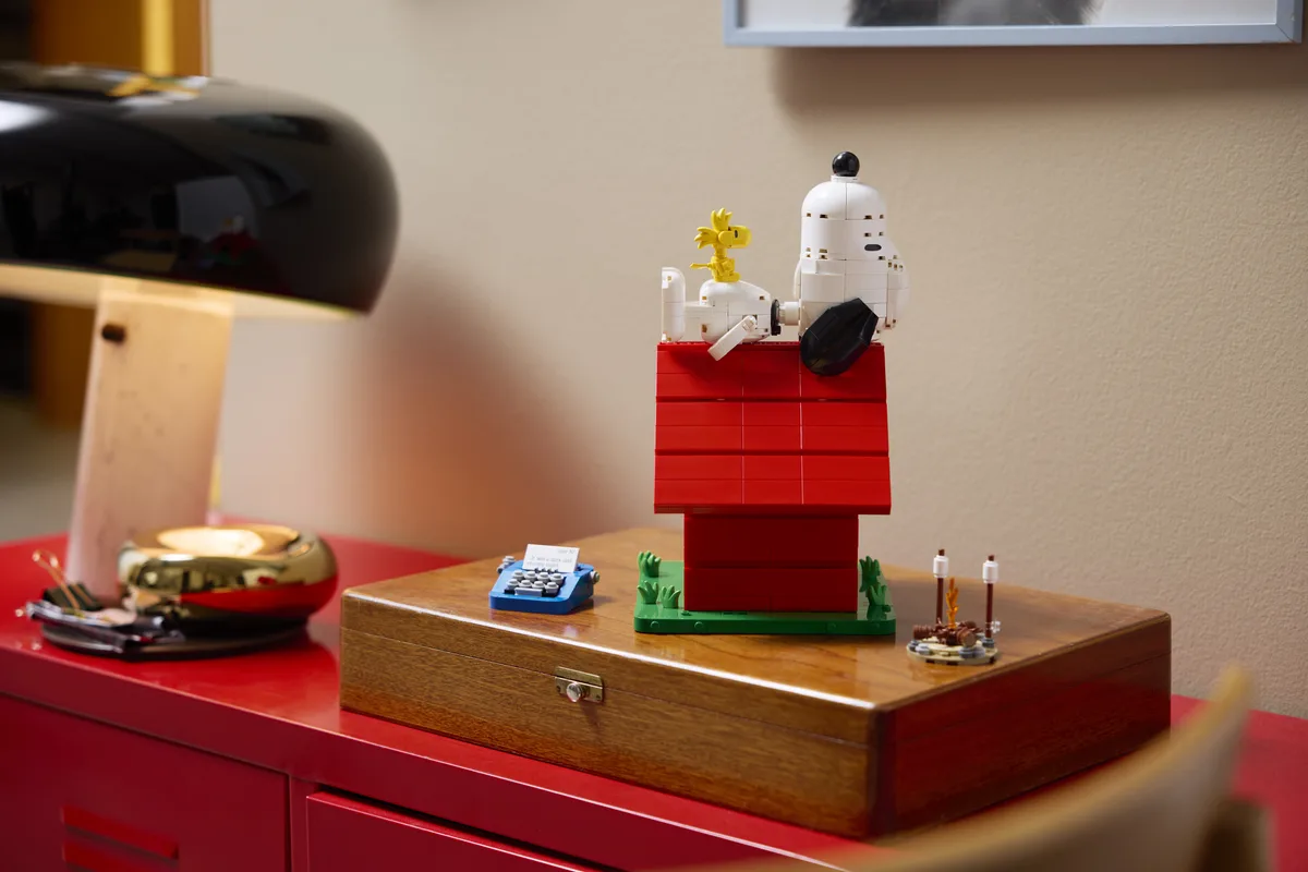 Snoopy and Woodstock sit on the roof of the red LEGO doghouse, next to a tiny typewriter, on a wooden box on a red sideboard